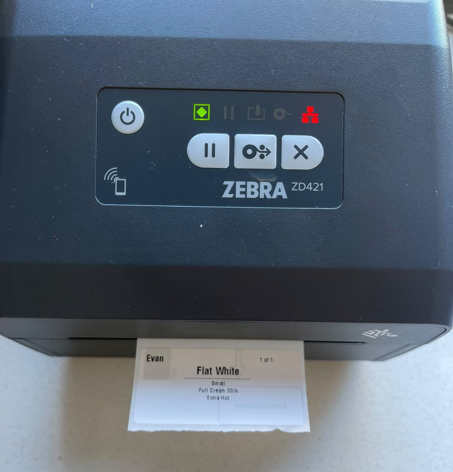 A Zebra ZD421 printer with a freshly printed sticker reading Flat White, small, full cream milk, extra hot